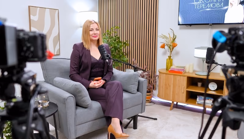 Eine Frau in einem lila Anzug sitzt auf einem grauen Sessel in einem Studio, während Mikrofone und Kameras sie aufnehmen.