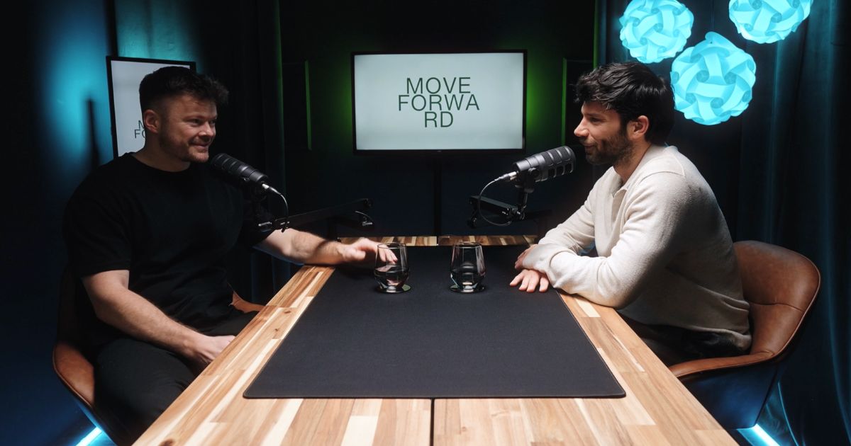 Zwei Männer sitzen an einem Holztisch mit Mikrofonen und Wassergläsern und führen ein Gespräch, im Hintergrund ein Bildschirm mit der Aufschrift 'MOVE FORWARD'.