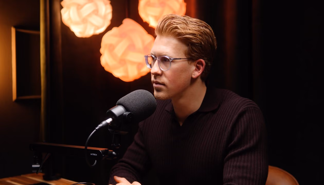 Ein junger Mann mit Brille spricht in ein Mikrofon vor drei orangefarbenen Lampen.