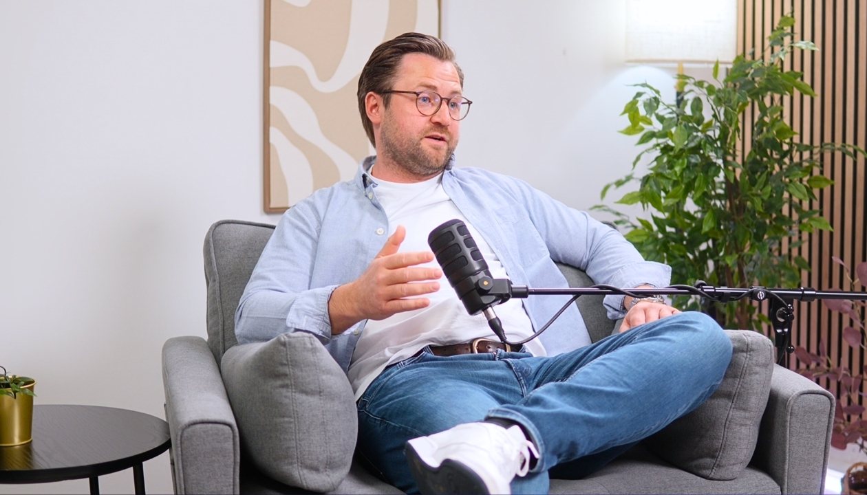 Mann in Jeans und Brille spricht sitzend auf grauem Sofa vor Mikrofon in modernem Raum.