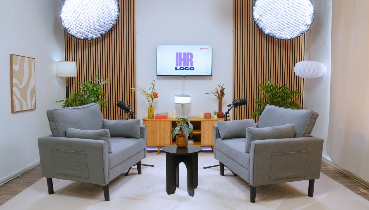 Zwei graue Sessel mit Mikrofonen vor TV und Pflanzen in einem modernen Podcast-Studio.