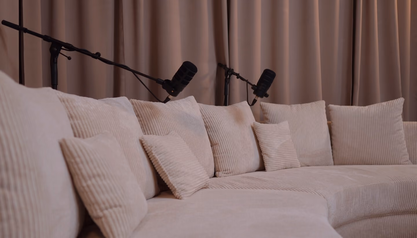 Ein geschwungenes, hellbeiges Sofa mit mehreren beigen Kissen vor einem beigefarbenen Vorhang, mit zwei schwarzen Mikrofonen, die auf das Sofa gerichtet sind.