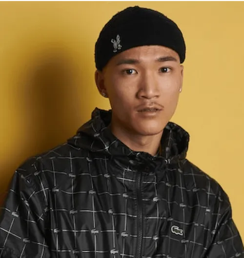 Person wearing a black patterned jacket and black beanie against a yellow background.