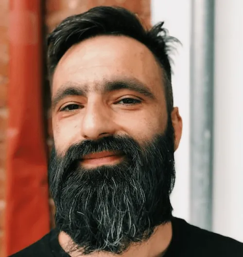 Person with a long beard and dark hair standing against a brick wall and metal pole.