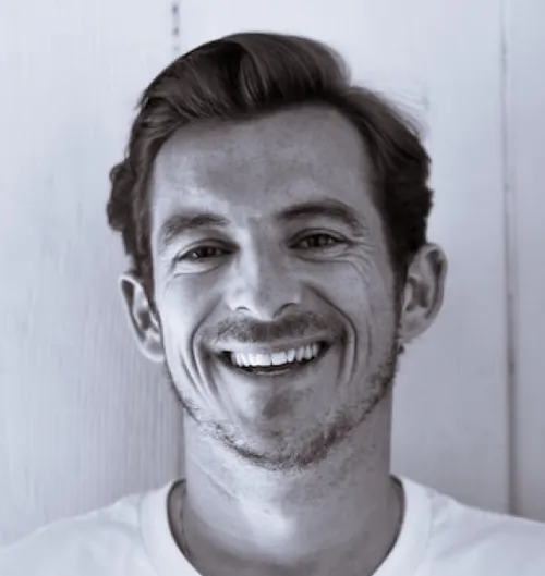 Person with short hair wearing a white shirt standing against a white wooden wall.
