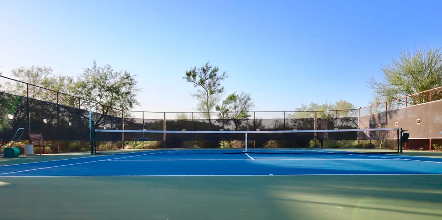 Empty outdoor tennis court with blue surface, net, and surrounding trees under clear sky.