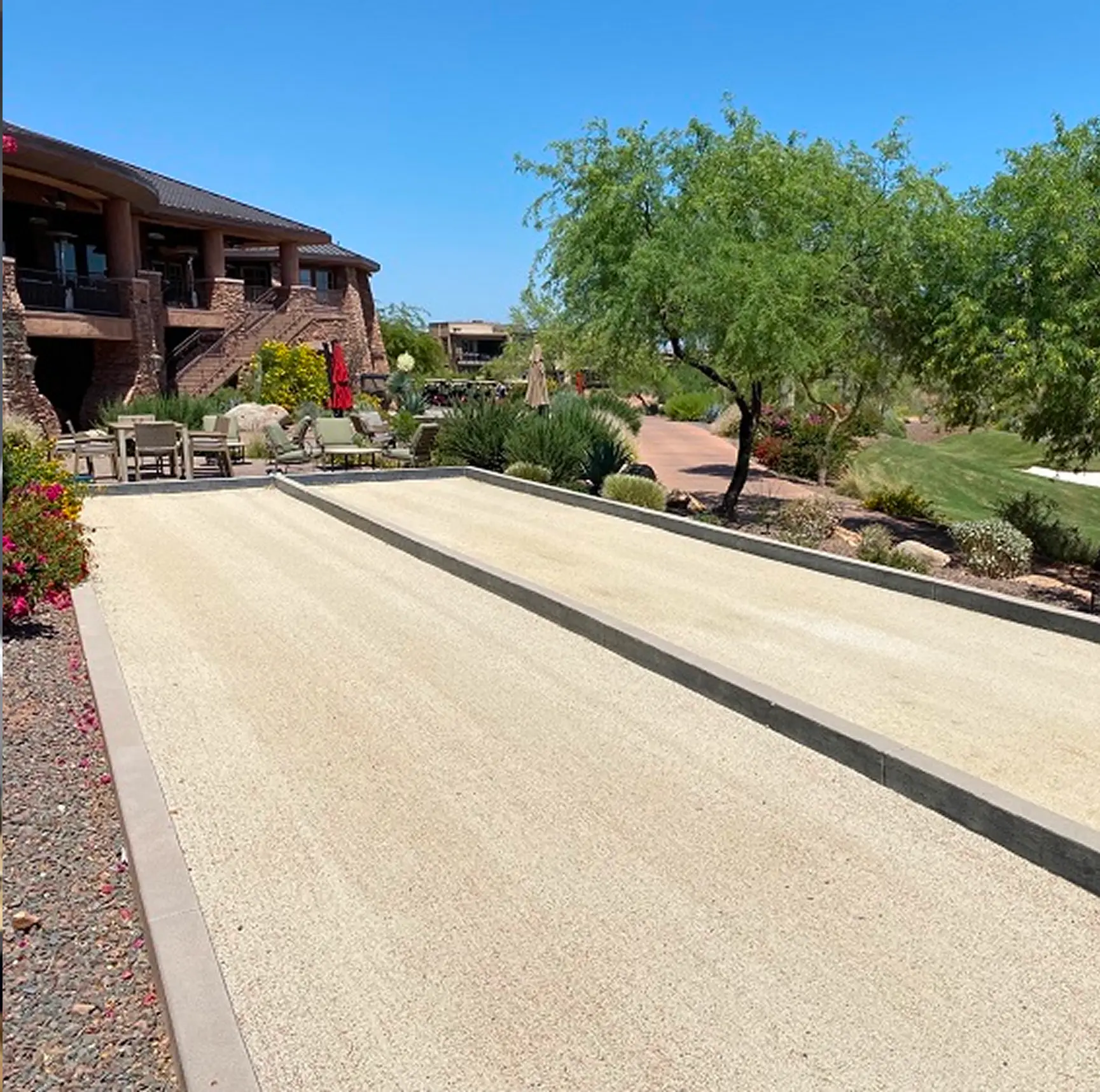 Empty outdoor bocce ball court with patio chairs, trees, and a stone building under clear blue sky.