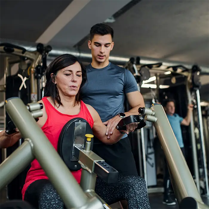 Woman in red tank top using gym machine with a male trainer assisting her arm movement.