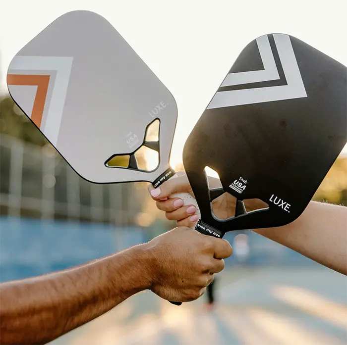 Two people holding black pickleball paddles with white designs, touching them together outdoors.