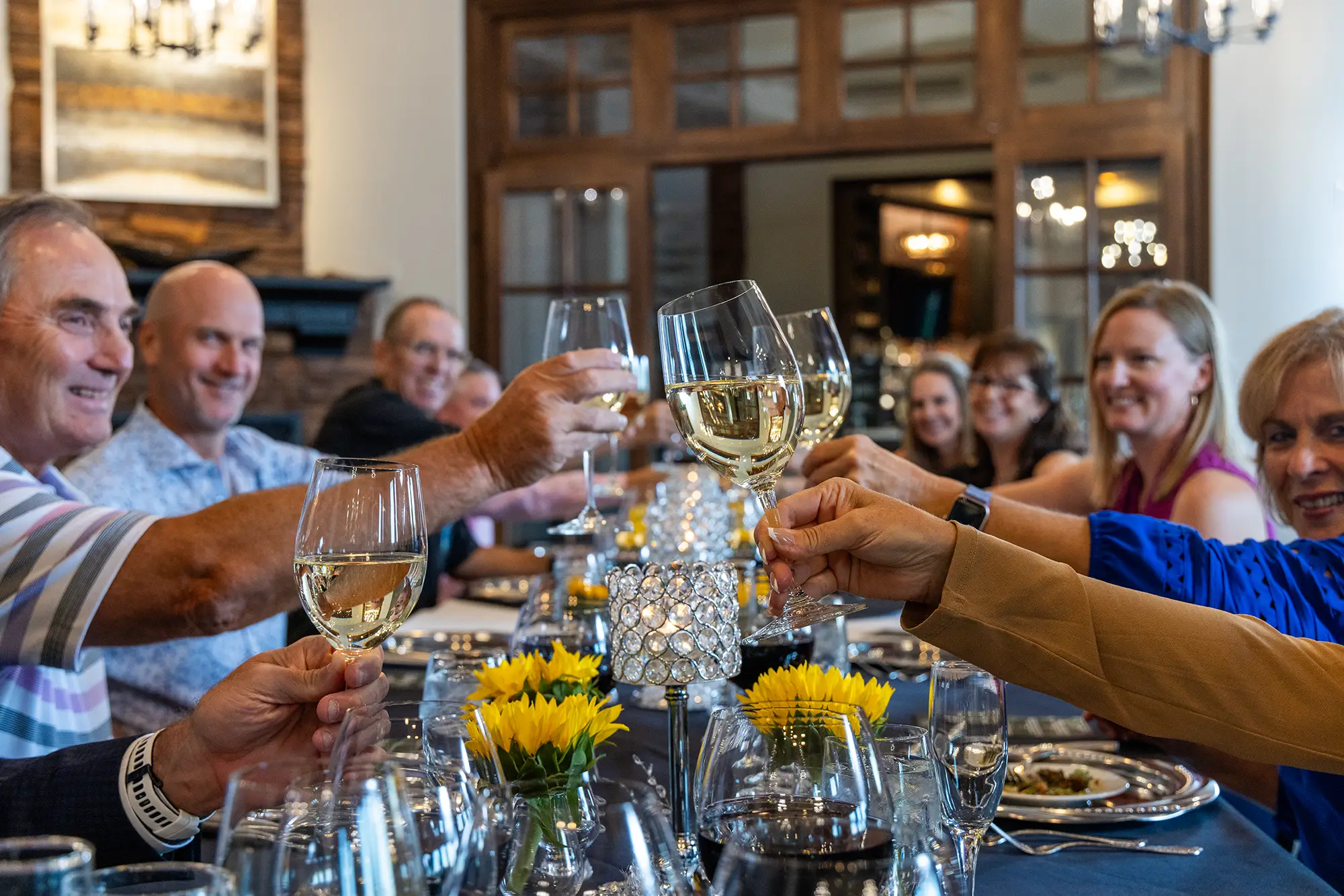 Group of people raising white wine glasses for a toast at a dining table with yellow flowers.