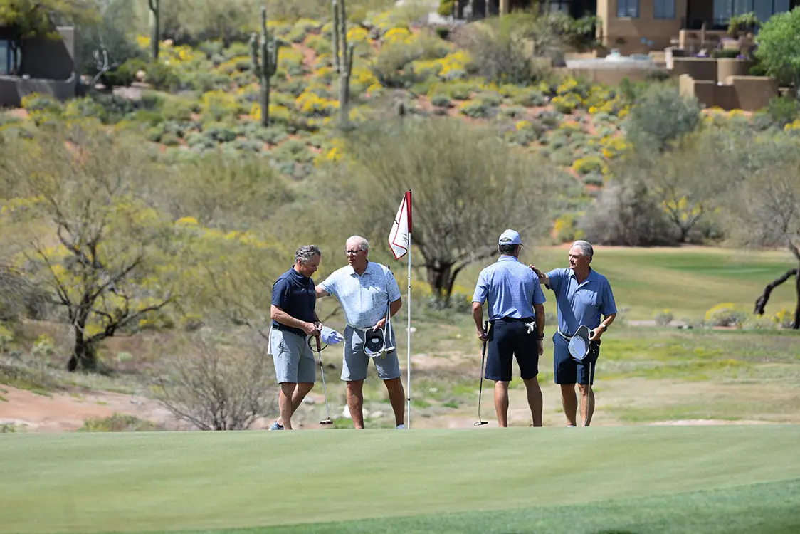 Four men standing near a golf hole on a green with desert plants in the background.