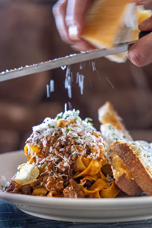 Hand grating cheese over a plate of pasta with meat sauce and two slices of garlic bread.