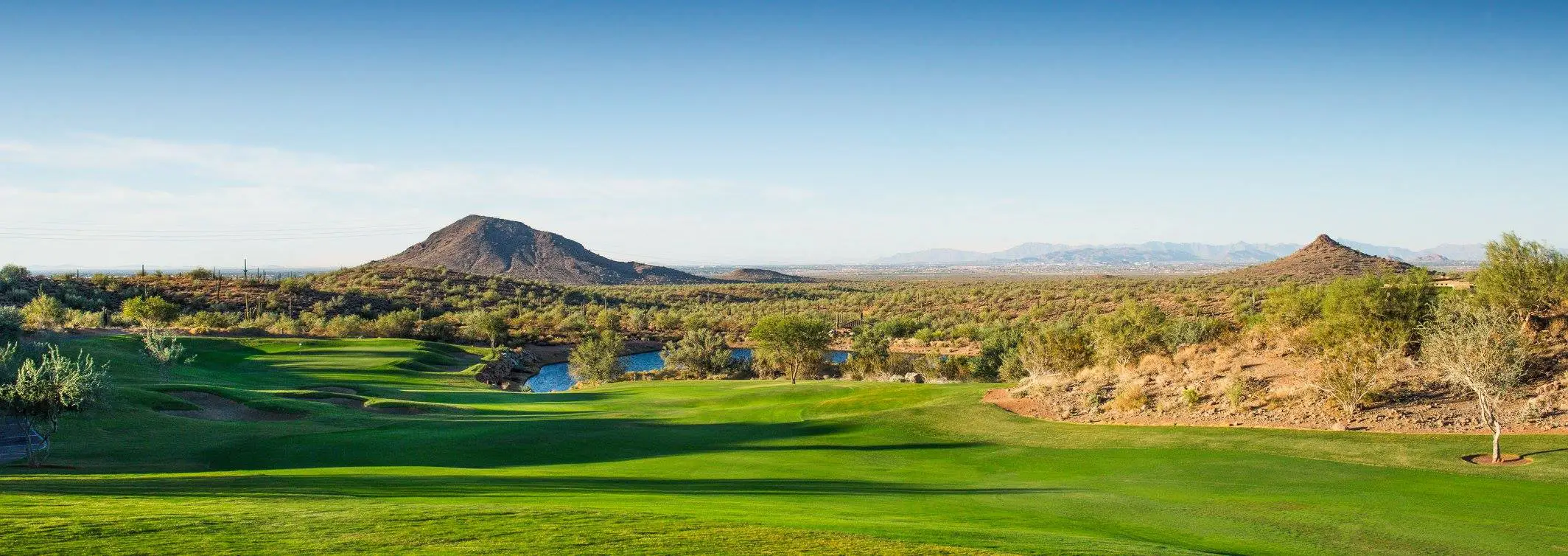 Panoramic view of a green golf course with desert hills and water hazards under a clear blue sky.