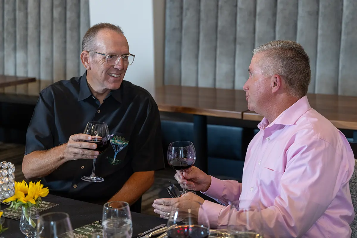 Two men sitting at a dining table, toasting with glasses of red wine.
