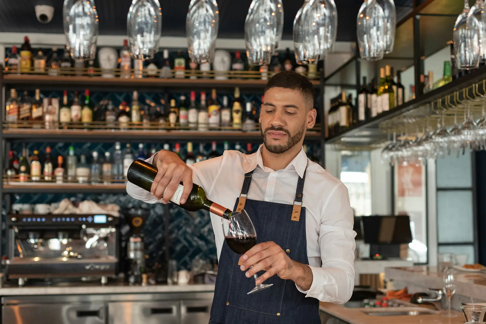 Bartender in white shirt and apron pouring red wine into a glass behind a bar.