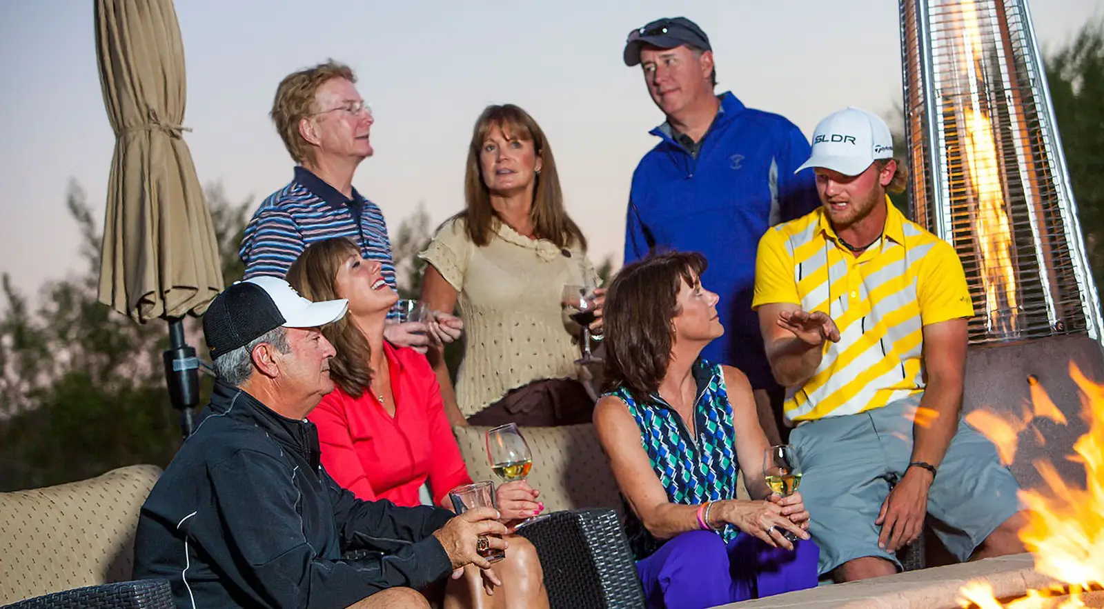 Group of six adults socializing outdoors around a fire pit at dusk with drinks and patio heater.
