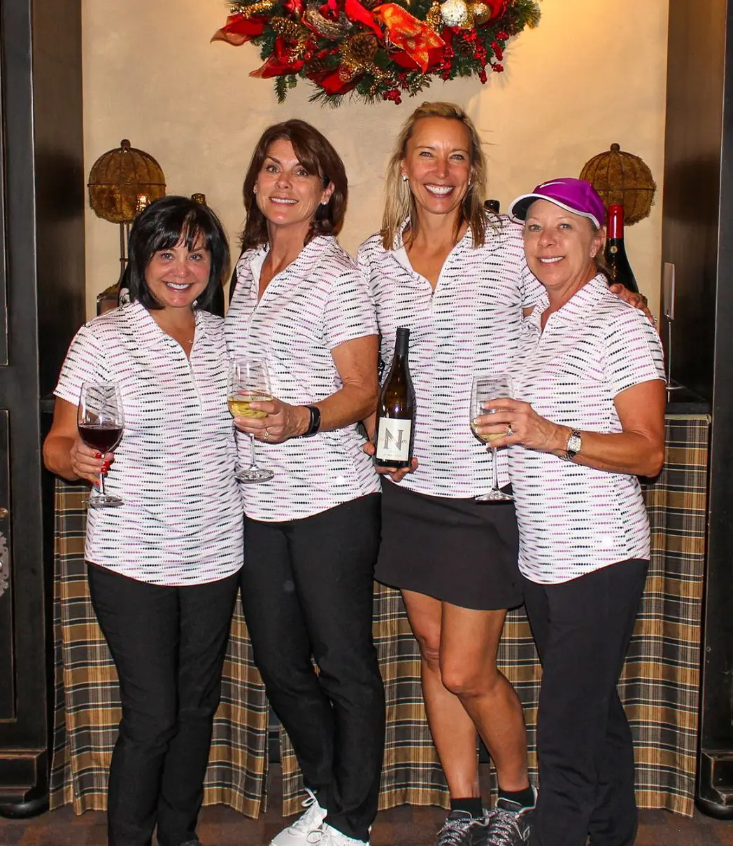 Four women in matching shirts holding wine glasses and a bottle, standing under a festive wreath.