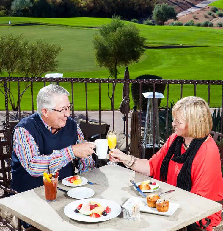 Older man and woman toasting drinks at outdoor table with breakfast overlooking golf course.