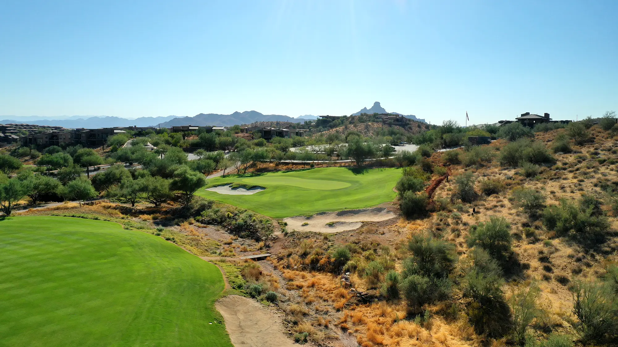 Golf course fairway and green surrounded by desert vegetation and distant mountain landscape.