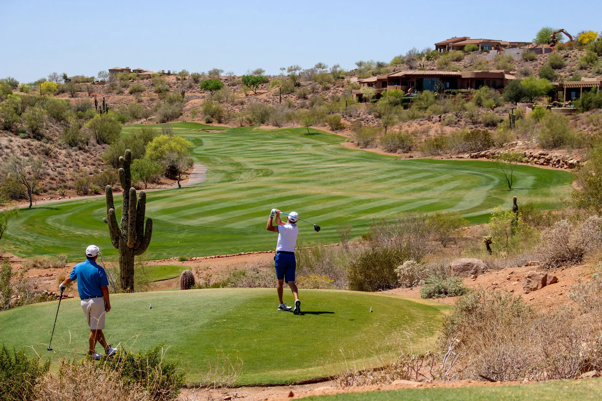 Two golfers on a tee box overlooking a green fairway with desert landscape and houses in background.