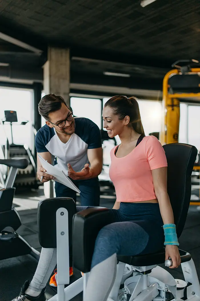 Trainer showing a workout plan to a smiling woman using leg press machine in gym.