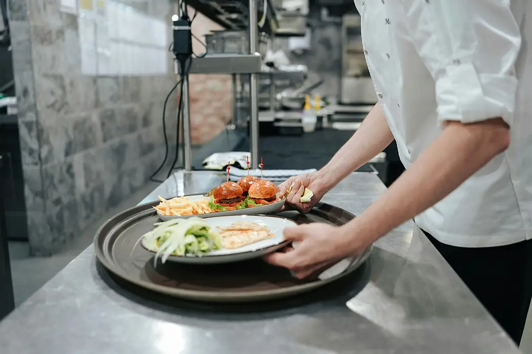 Chef placing plates with burgers, fries, and salad on a large tray in a kitchen.