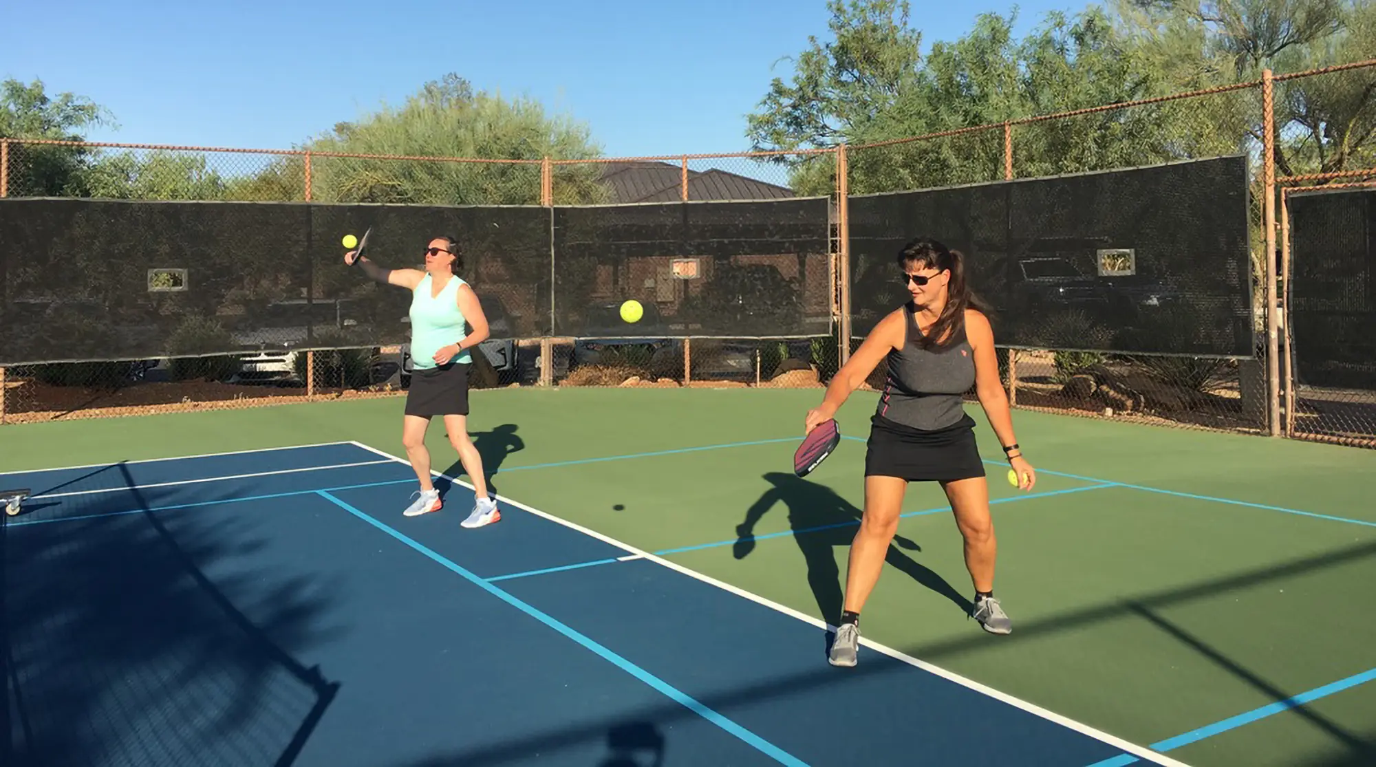 Two women playing pickleball on an outdoor court under a clear sky with paddles and balls mid-air.