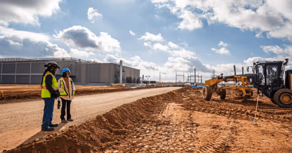Two construction workers in safety vests on a dirt road at an active data center construction site