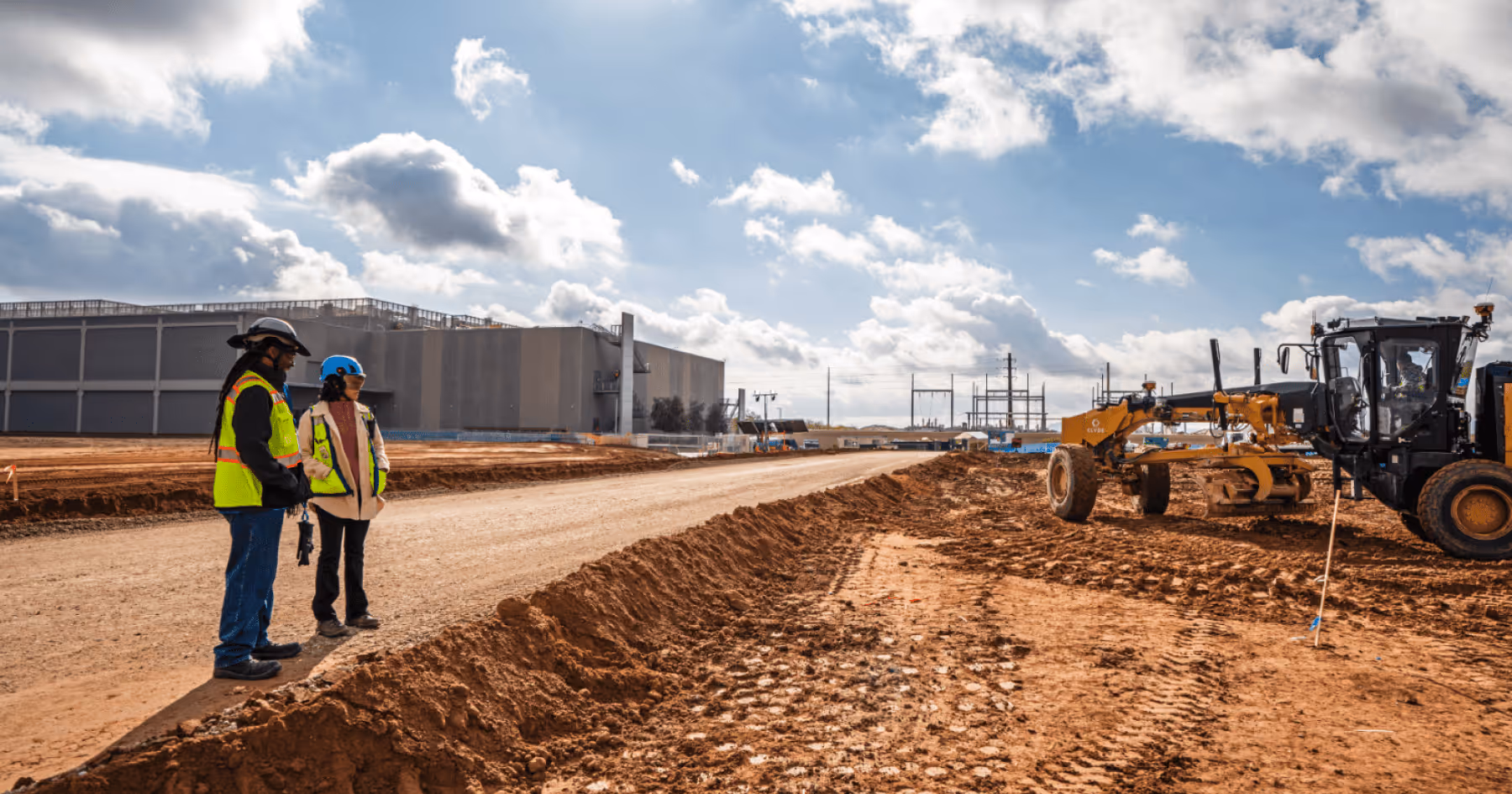 Two construction workers in safety vests on a dirt road at an active data center construction site