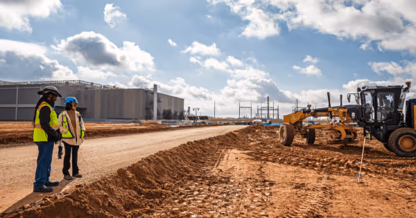 Two construction workers in safety vests standing on a dirt road next to a grading machine