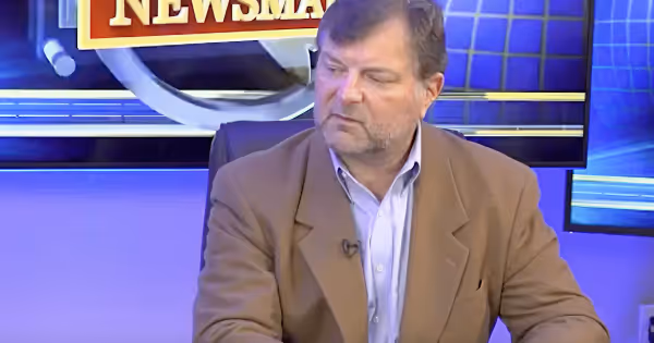Man in a tan blazer seated at a news desk with a Newsmax TV backdrop