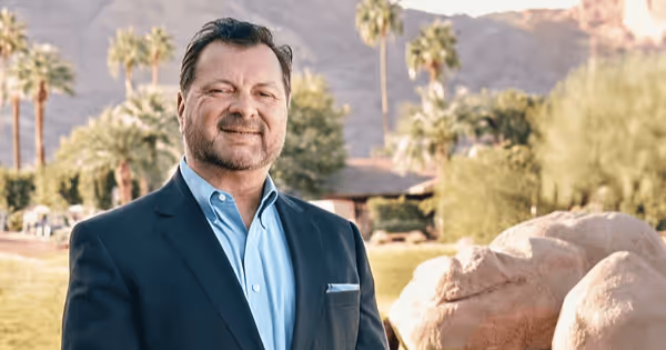 Man in a blue sport coat standing outdoors with palm trees and boulders in the background