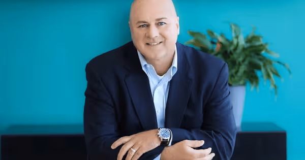 Headshot of a bald man in a blue blazer smiling against a teal background with a plant