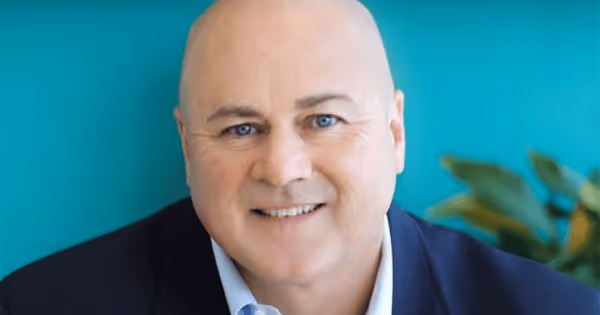 Headshot of a bald man in a blue blazer smiling against a teal background with a plant