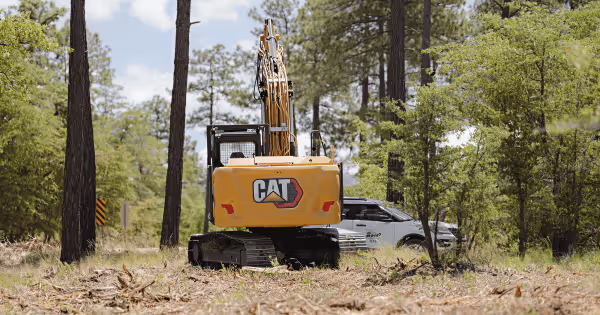 A yellow CAT excavator clearing trees in a forest setting