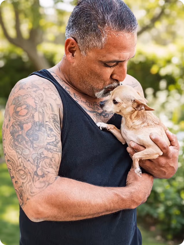 Man with tattoos wearing a black tank top gently holding and kissing a small beige dog outdoors.