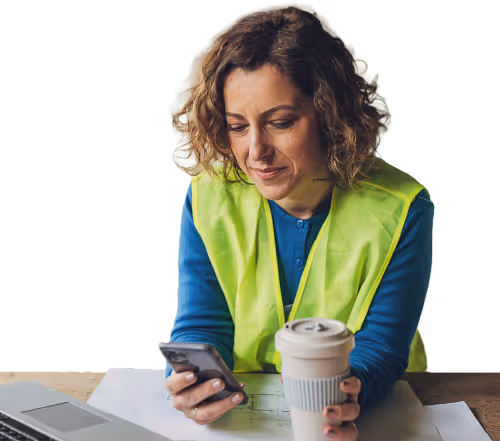 Woman wearing a yellow safety vest looking at her phone while holding a reusable coffee cup, sitting at a table with papers and a laptop.