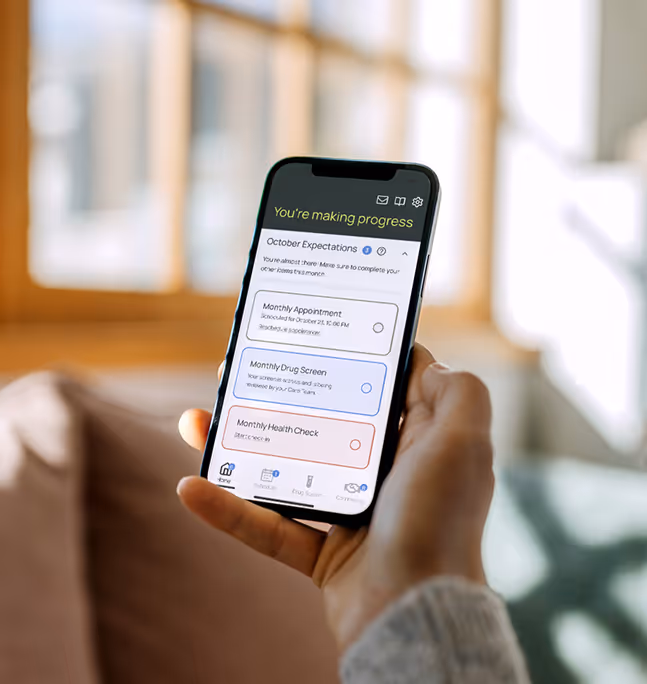 Hand holding a smartphone displaying a progress tracking app with October tasks including monthly appointment, drug screen, and health check.