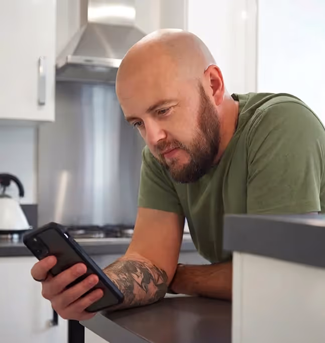 Bald man with beard and tattoo on forearm looking at smartphone while leaning on kitchen counter.