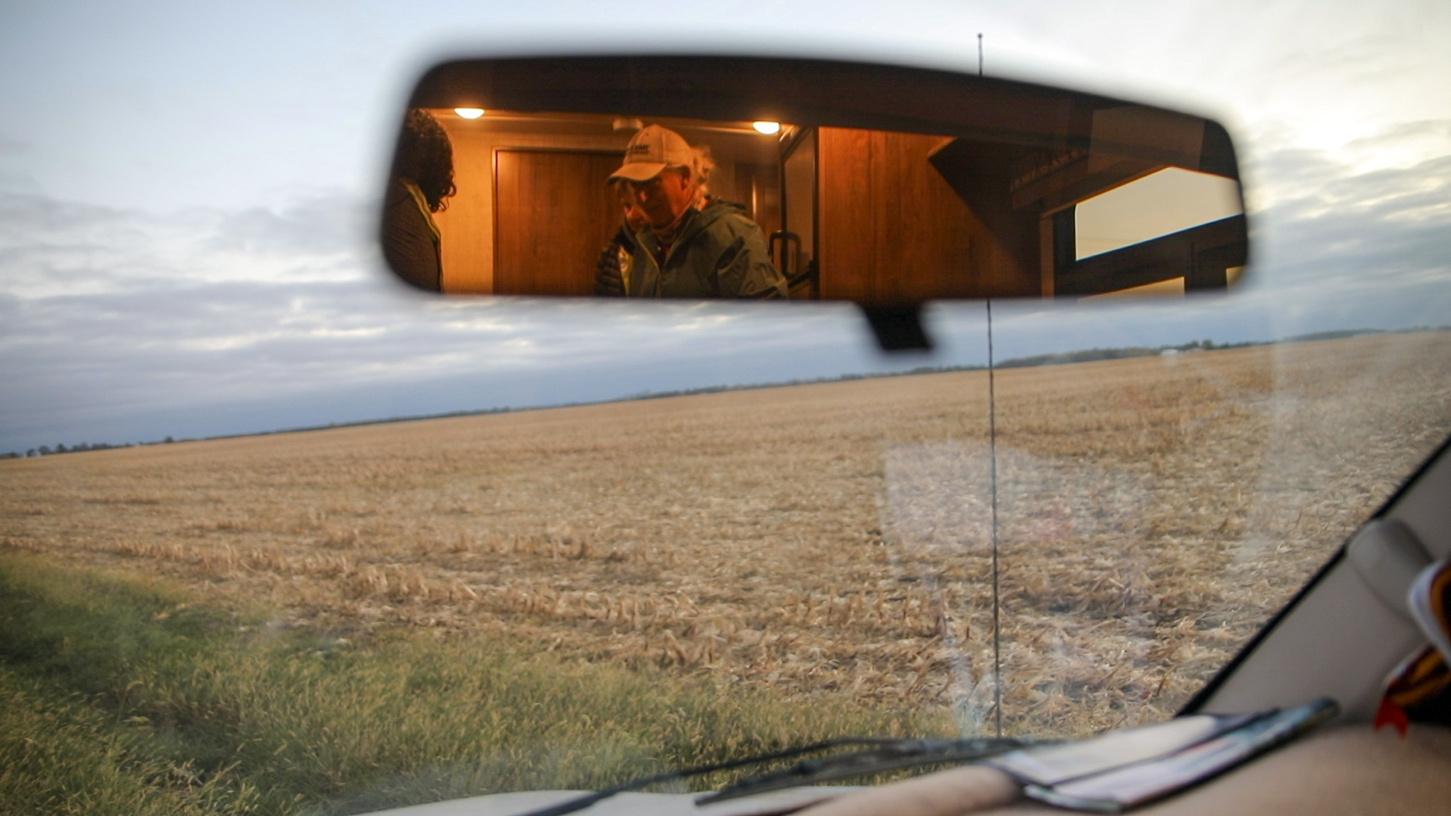 View through a vehicle windshield of a vast harvested field under a cloudy sky, with three people reflected in the rearview mirror inside a wooden room.