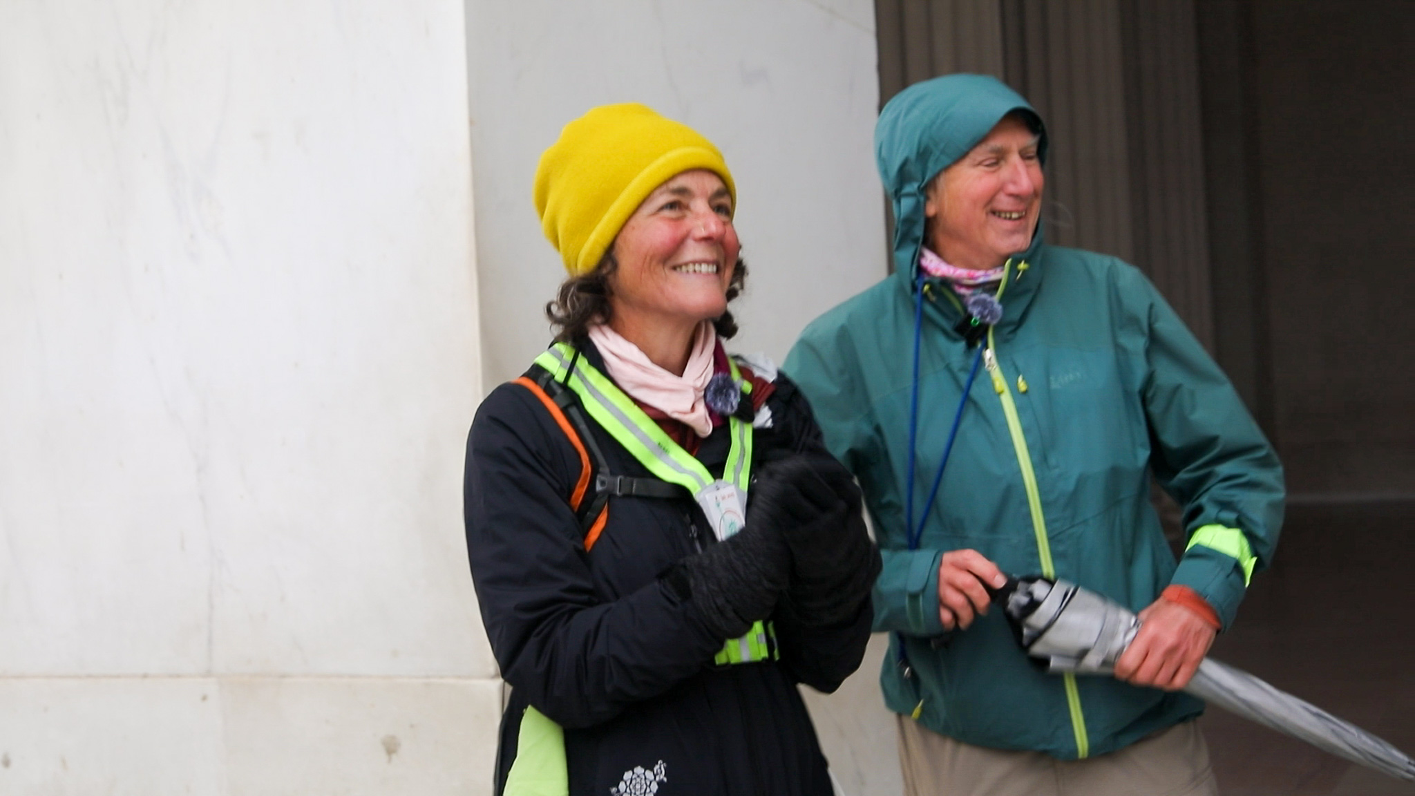 Two people dressed in outdoor gear smiling, one wearing a yellow beanie and the other holding a folded umbrella.