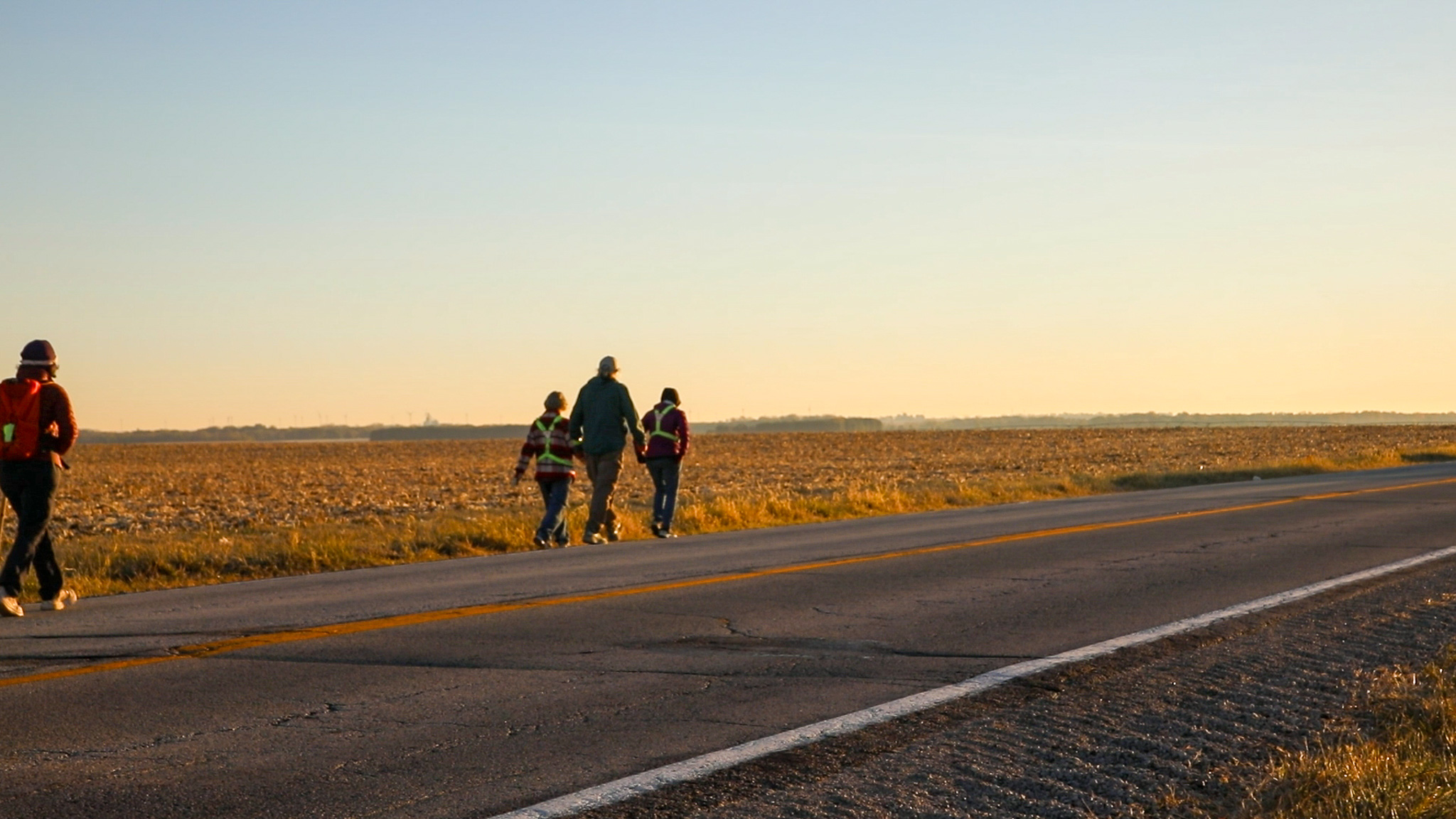 Four people walking along a rural road next to a harvested field at sunset.