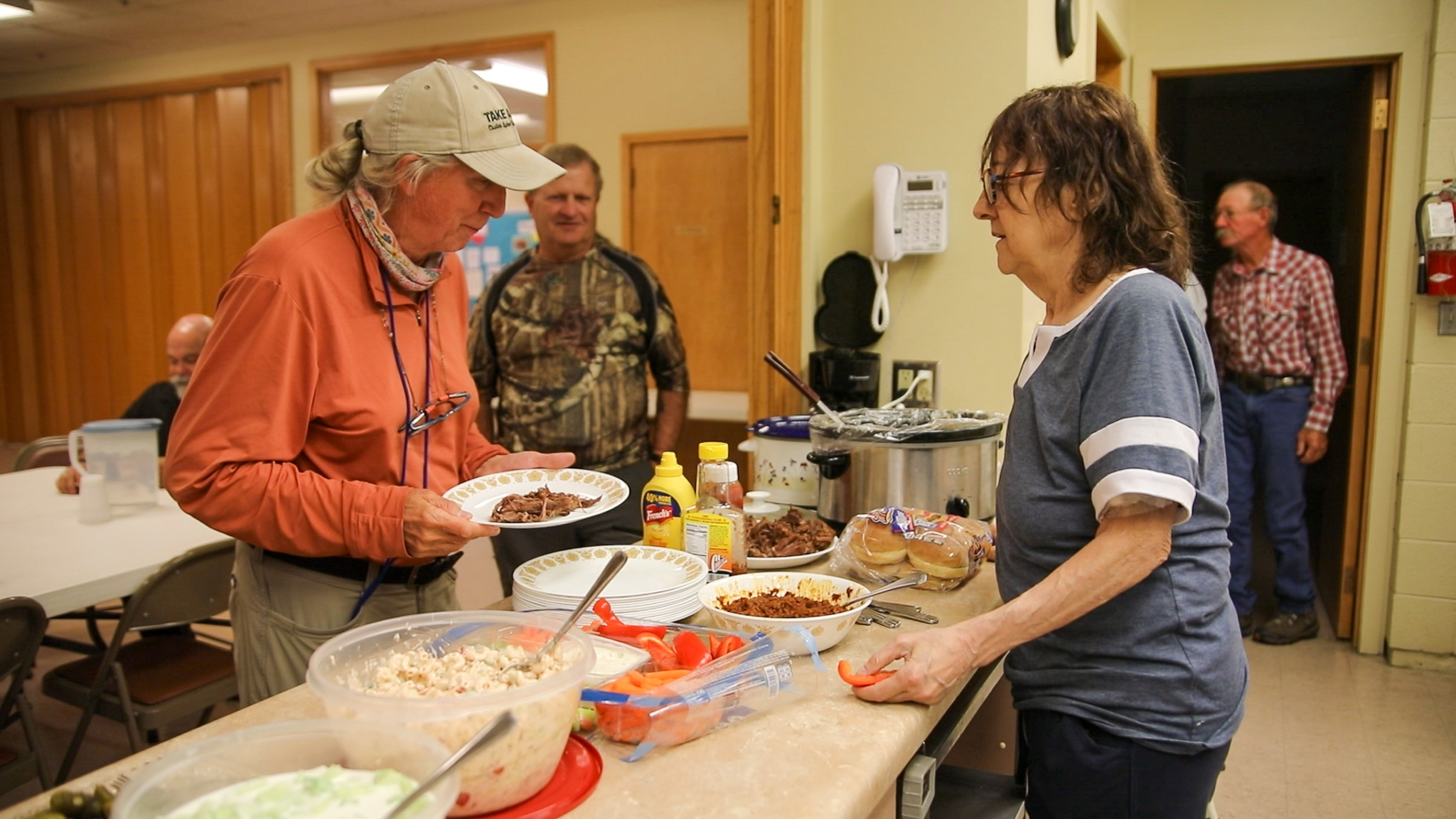 Two women serving food at a buffet table with bowls of salad, vegetables, and condiments in a community dining room.