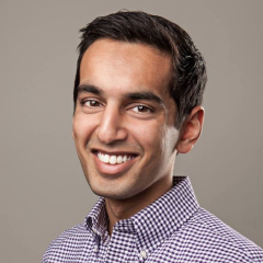 Smiling man with short dark hair wearing a purple and white checkered shirt against a plain gray background.