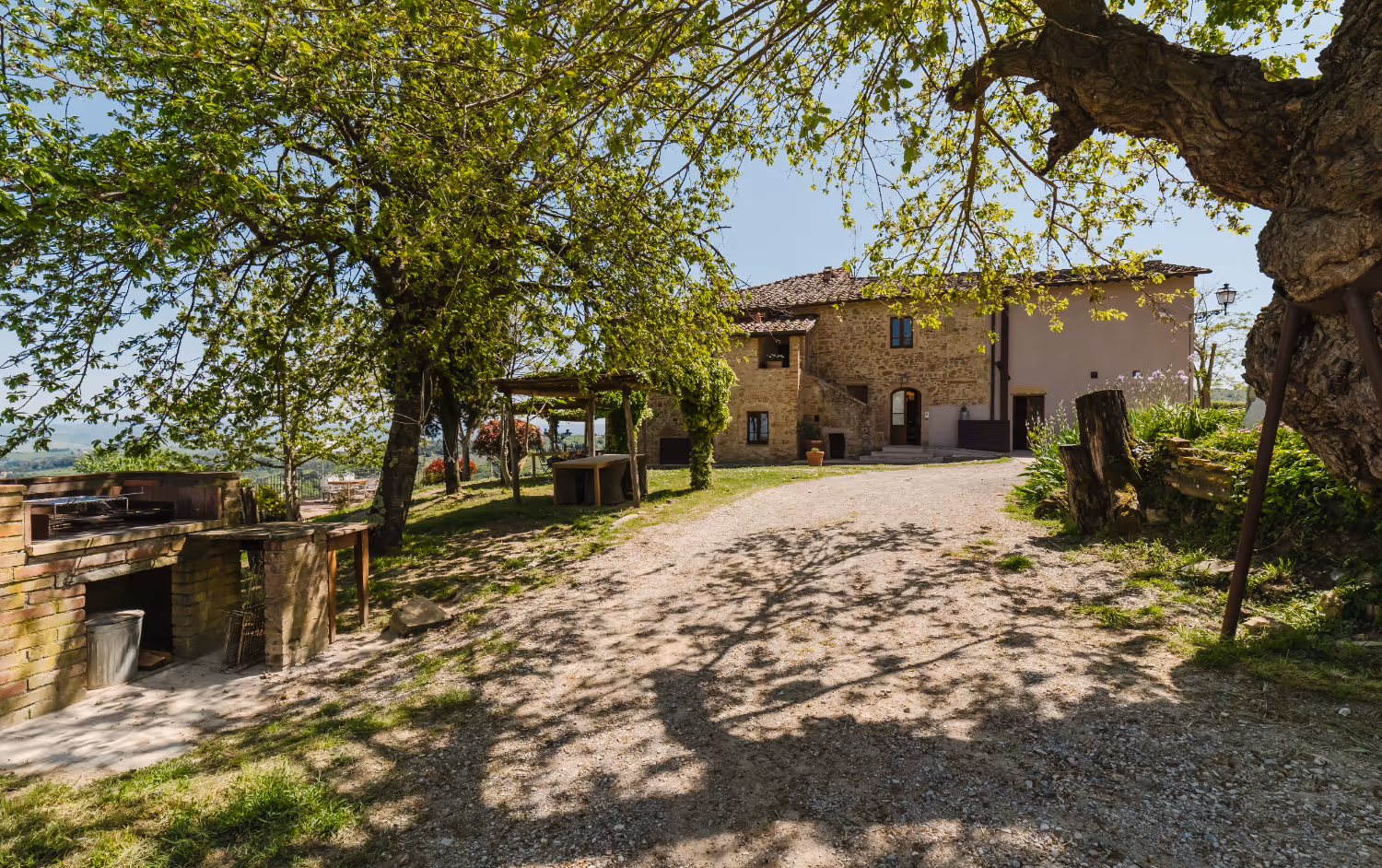 Stone house with tile roof in countryside surrounded by trees and a gravel driveway.