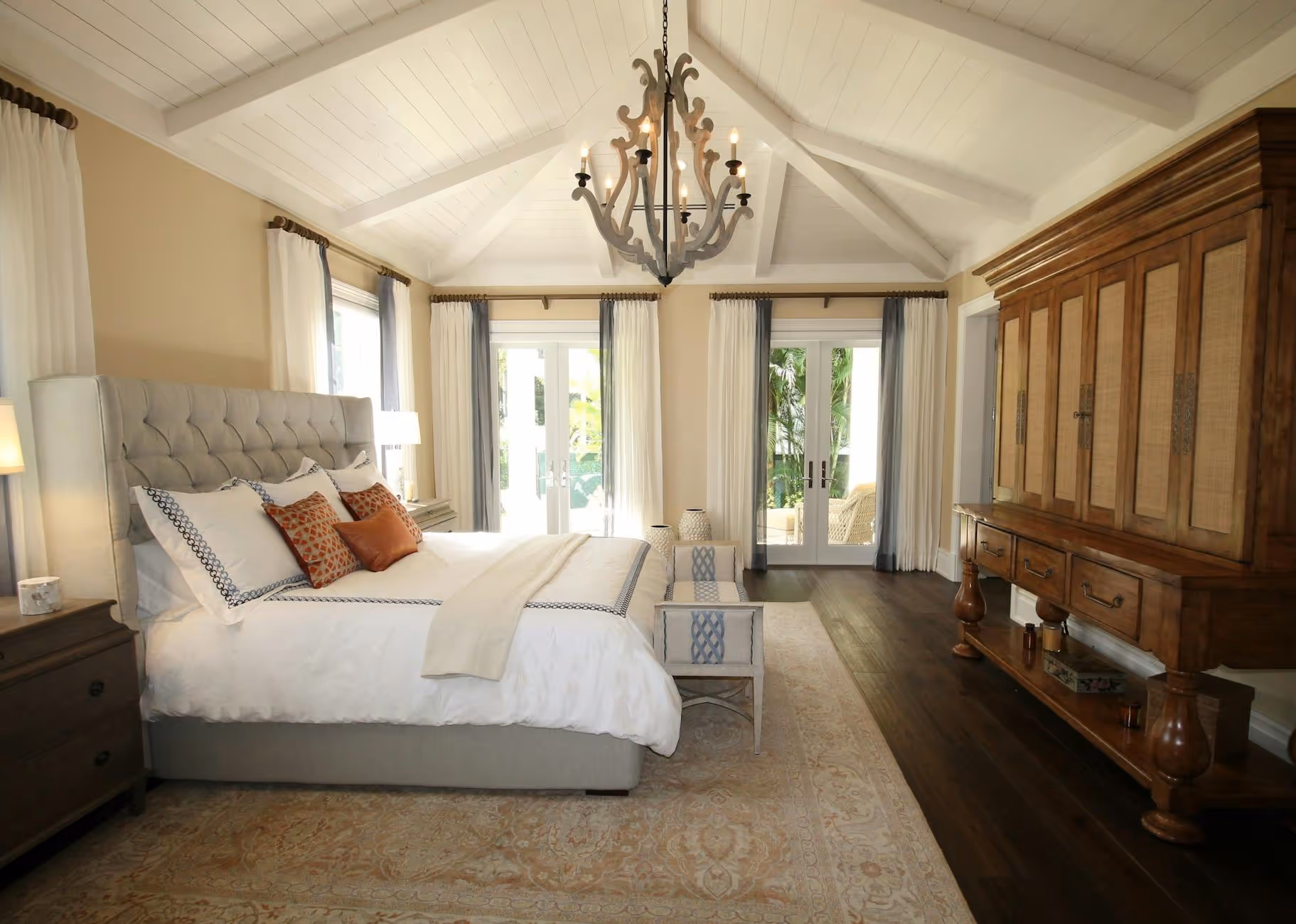 Bright bedroom with a tufted bed, white bedding, brown wooden furniture, and glass doors opening outside.