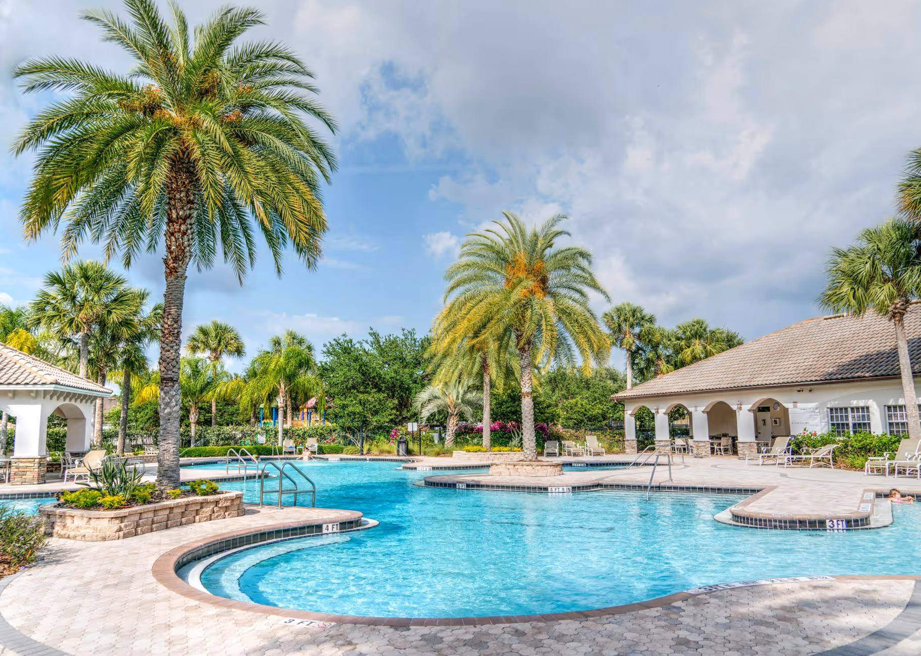 Bright outdoor swimming pool surrounded by palm trees and sun loungers under a partly cloudy sky.