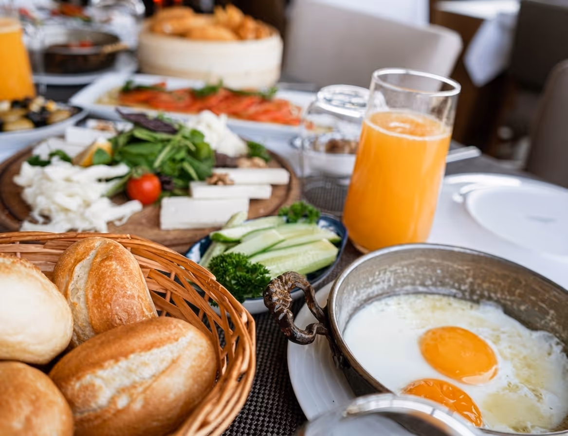 Breakfast table with basket of rolls, fried eggs in pan, sliced cucumber, orange juice, cheese, and greens.