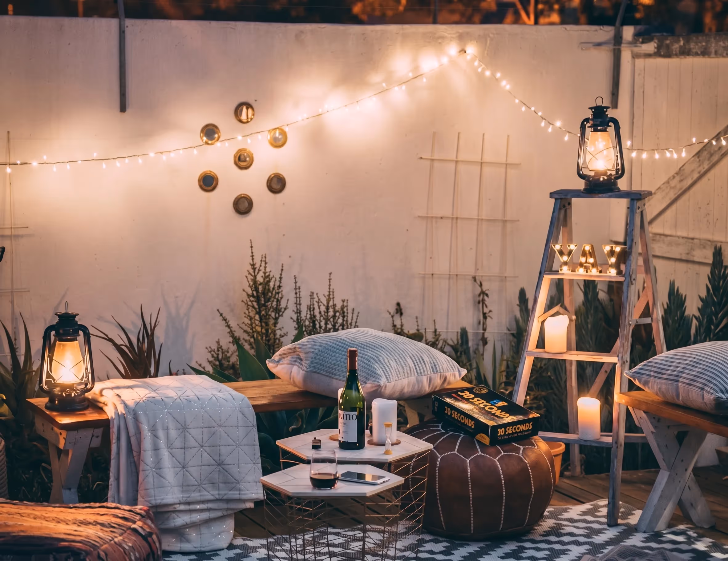 Cozy outdoor seating area at dusk with cushions, lanterns, wine, and a 30 Seconds board game.