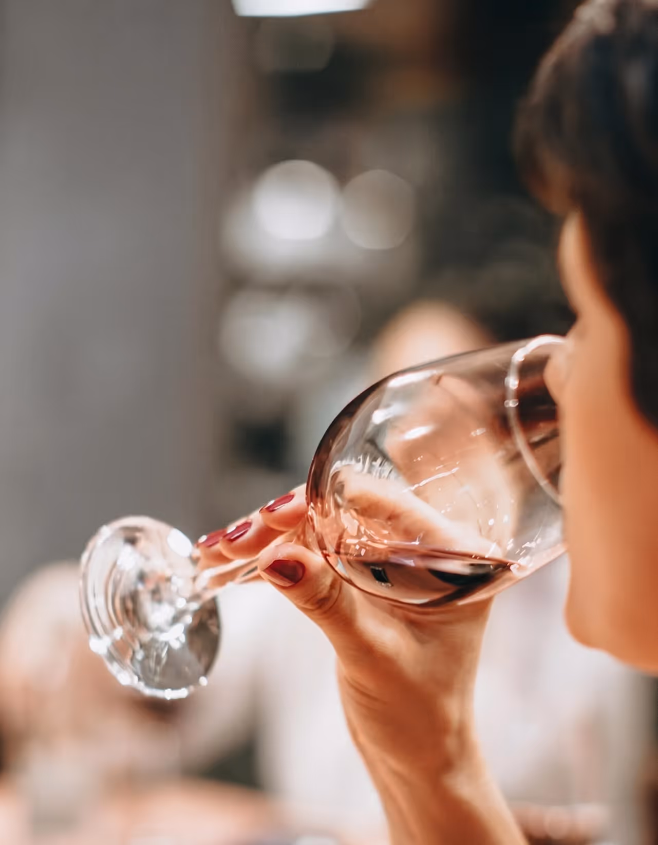 Person holding a wine glass with red wine near their mouth in a social setting.
